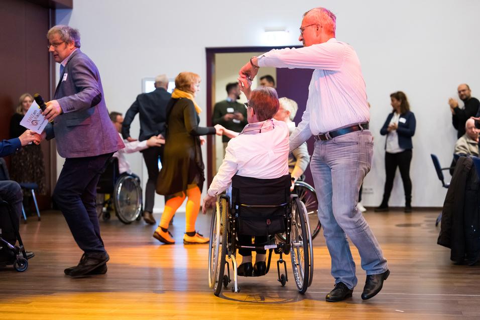 Tanzende Menschen in einem Raum, darunter eine Person im Rollstuhl, die von einem Mann angeführt wird.